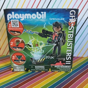 May include: Playmobil Ghostbusters II Spengler figure with a proton pack and a ghost trap. The figure is holding a proton pack and is standing in front of a ghost trap. The box has a warning label for choking hazards. The box also has a QR code and the text "playmogram.playmobil.com".