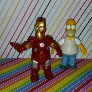 Op de afbeelding: Twee beeldjes: een is een rode en gouden Iron Man met een Homer Simpson-hoofd, de andere is Homer Simpson in een wit shirt, een blauwe broek en een gele huid. Ze staan op een kleurrijk gestreept oppervlak.
