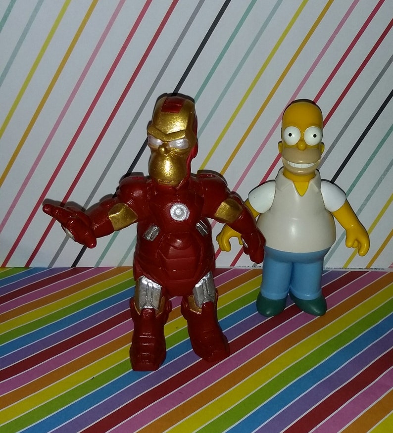 Op de afbeelding: Twee beeldjes: een is een rode en gouden Iron Man met een Homer Simpson-hoofd, de andere is Homer Simpson in een wit shirt, een blauwe broek en een gele huid. Ze staan op een kleurrijk gestreept oppervlak.