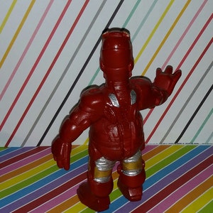 Op de afbeelding: Een rode Iron Man actiefiguur met zilveren en gouden accenten. De figuur staat met &eacute;&eacute;n arm omhoog. Het speelgoed staat op een kleurrijke gestreepte achtergrond.