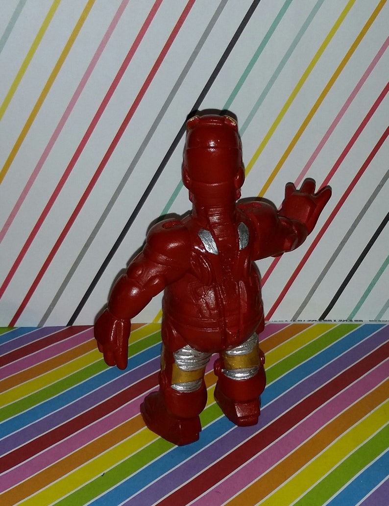 Op de afbeelding: Een rode Iron Man actiefiguur met zilveren en gouden accenten. De figuur staat met &eacute;&eacute;n arm omhoog. Het speelgoed staat op een kleurrijke gestreepte achtergrond.
