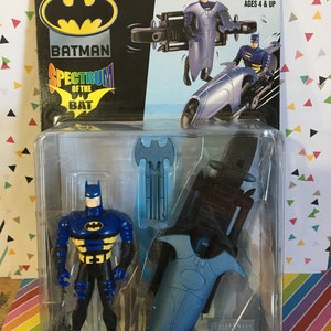 May include: A Batman action figure in a plastic package. The figure is blue and black with a yellow bat symbol. The package also includes a blue weapon and the text "GAMMA BLAST BATMAN". The packaging includes the text "Spectrum of the Bat".