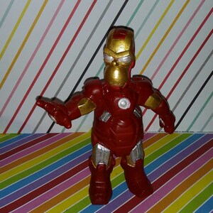Op de afbeelding: Een rood en goud beeldje van een personage dat lijkt op Homer Simpson in een Iron Man-pak. De figuur heeft een gouden helm en accenten, met zilveren details op de benen en armen. De achtergrond is een kleurrijk strepenpatroon.