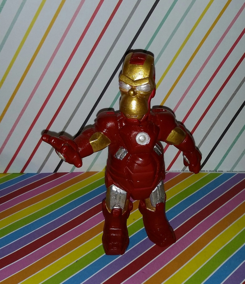 Op de afbeelding: Een rood en goud beeldje van een personage dat lijkt op Homer Simpson in een Iron Man-pak. De figuur heeft een gouden helm en accenten, met zilveren details op de benen en armen. De achtergrond is een kleurrijk strepenpatroon.
