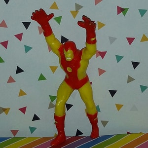 K&ouml;nnte beinhalten: Eine rote und gelbe Plastik-Actionfigur eines Superhelden mit erhobenen Armen. Die Figur steht auf einer bunten gestreiften Oberfl&auml;che.