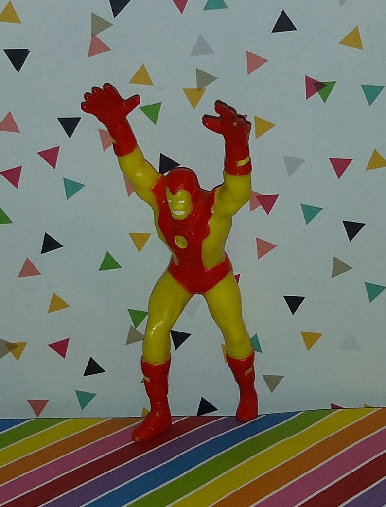 K&ouml;nnte beinhalten: Eine rote und gelbe Plastik-Actionfigur eines Superhelden mit erhobenen Armen. Die Figur steht auf einer bunten gestreiften Oberfl&auml;che.
