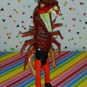 May include: A red and black action figure with a scorpion-like head and tail. The figure is wearing black pants and red boots.