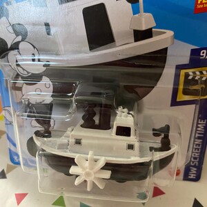 Puede incluir: Un barco de juguete blanco y negro con un personaje de Mickey Mouse en el lateral. El barco tiene una h&eacute;lice y una peque&ntilde;a bandera. El juguete est&aacute; empaquetado en un bl&iacute;ster de pl&aacute;stico transparente con fondo azul. El embalaje incluye el texto "HW SCREEN TIME".