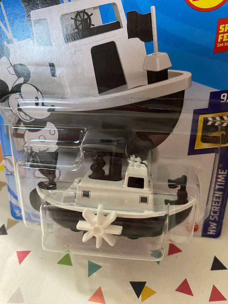 Puede incluir: Un barco de juguete blanco y negro con un personaje de Mickey Mouse en el lateral. El barco tiene una h&eacute;lice y una peque&ntilde;a bandera. El juguete est&aacute; empaquetado en un bl&iacute;ster de pl&aacute;stico transparente con fondo azul. El embalaje incluye el texto "HW SCREEN TIME".