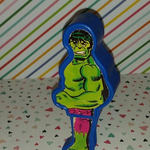 May include: A blue plastic figure of the Incredible Hulk. The figure is in a standing pose with his arms crossed. The Hulk is wearing purple pants and has a green and yellow comic book style design.