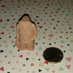 May include: A small, beige figurine of an alien with large eyes and a thin body. The figurine is sitting with its hands clasped in front of it. The figurine is about the size of a quarter.