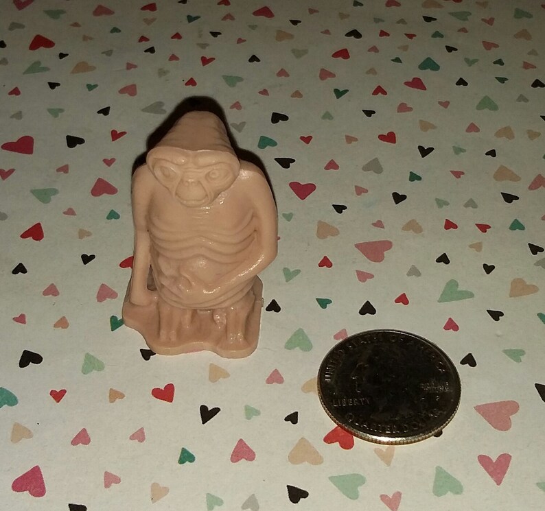 May include: A small, beige figurine of an alien with large eyes and a thin body. The figurine is sitting with its hands clasped in front of it. The figurine is about the size of a quarter.