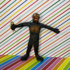 May include: A vintage gorilla toy with a brown face and chest, black fur, and outstretched arms, holding a small doll. The toy is set against a colorful striped background.