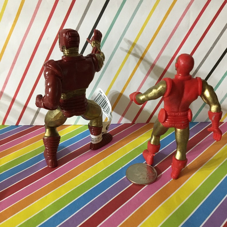 May include: Two red and gold plastic action figures of Iron Man. The figures are posed in a dynamic stance. The figure on the left is holding a weapon in its right hand. The figure on the right is holding a weapon in its left hand.