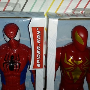 May include: Two action figures in their packaging. The figure on the left is Spider-Man, wearing a red and blue suit with a black spider logo on the chest. The figure on the right is Iron Spider, wearing a red suit with a gold spider logo on the chest. The packaging is white with red text that says "SPIDER-MAN" and "IRON SPIDER". The packaging also includes the text "AGES 4+".