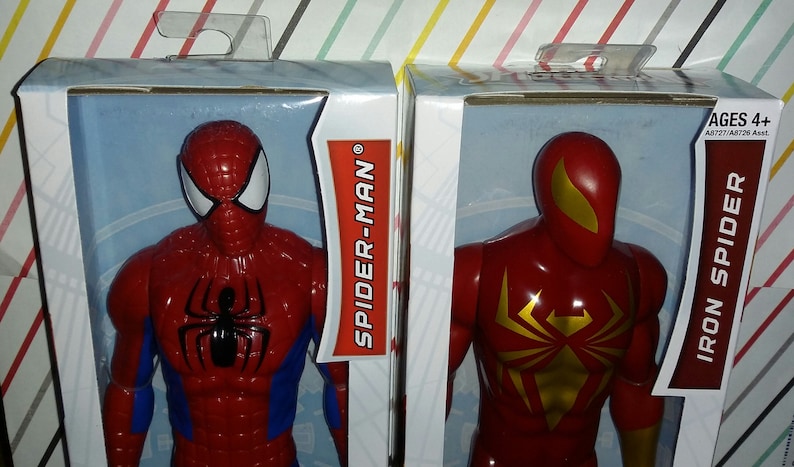 May include: Two action figures in their packaging. The figure on the left is Spider-Man, wearing a red and blue suit with a black spider logo on the chest. The figure on the right is Iron Spider, wearing a red suit with a gold spider logo on the chest. The packaging is white with red text that says "SPIDER-MAN" and "IRON SPIDER". The packaging also includes the text "AGES 4+".