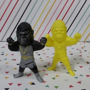 May include: Two toy gorilla figurines. One is black with a gold chain and gray outfit. The other is yellow with a gold chain.