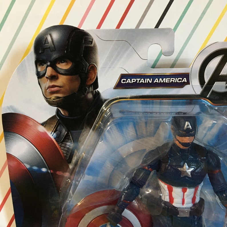 May include: A plastic action figure of Captain America in his blue and red costume, holding his shield. The figure is packaged in a clear plastic blister pack with a blue and white background. The packaging also features a larger image of Captain America's face and the text "Captain America".