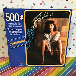 May include: A blue and yellow box for a 500 piece jigsaw puzzle featuring a photo of a woman from the movie Flashdance. The box has the text "Flashdance" and "Blockbuster" on it.