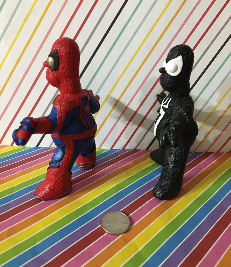 Peut inclure: Deux petites figurines peintes &agrave; la main de Spider-Man et Venom. Spider-Man est rouge et bleu avec des d&eacute;tails noirs. Venom est noir avec des yeux blancs. Les figurines sont debout sur un fond ray&eacute; color&eacute;.