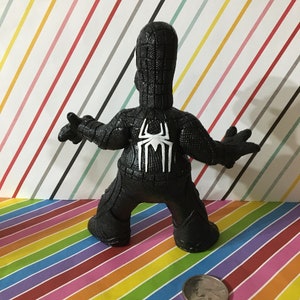Peut inclure: figurine en plastique noire et blanche d'un super-h&eacute;ros portant un costume noir avec un motif de toile d'araign&eacute;e au dos. La figurine est debout sur un fond ray&eacute;.