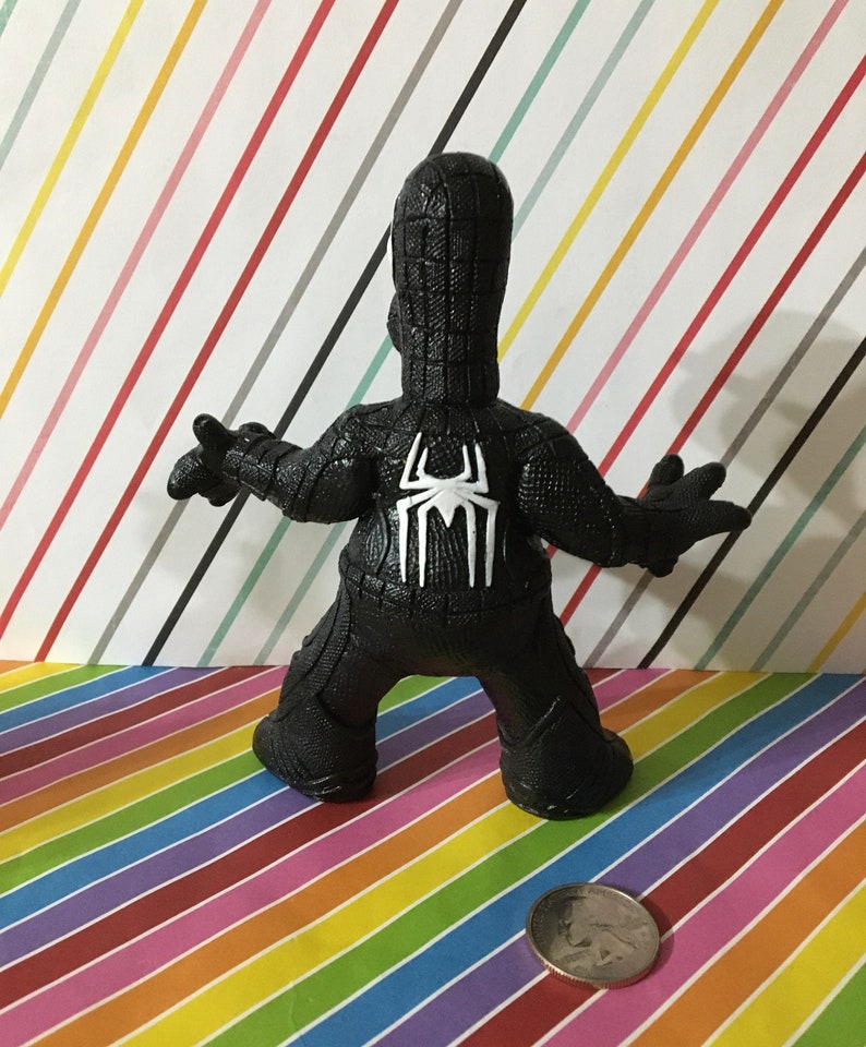 Peut inclure: figurine en plastique noire et blanche d'un super-h&eacute;ros portant un costume noir avec un motif de toile d'araign&eacute;e au dos. La figurine est debout sur un fond ray&eacute;.