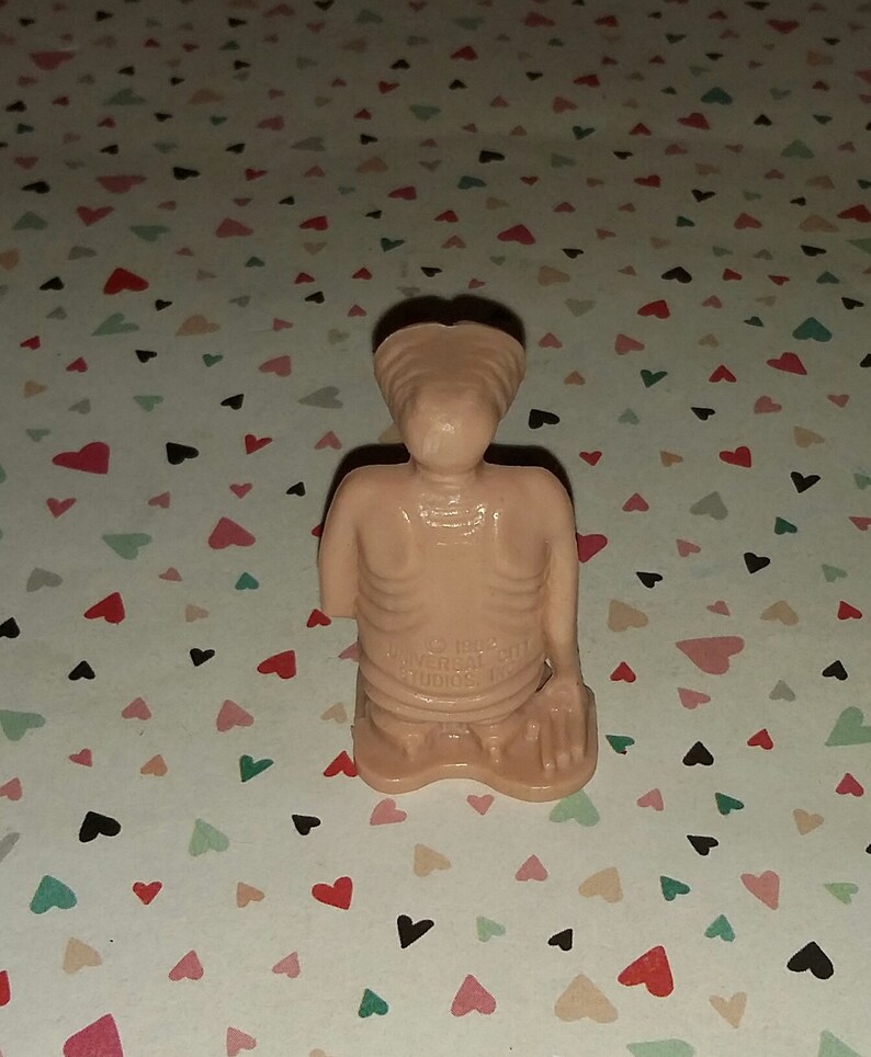 May include: A small, beige plastic figurine of an alien with a large head and a thin body. The figurine is in a kneeling position with its hands clasped together. The figurine is made by Universal City Studios.