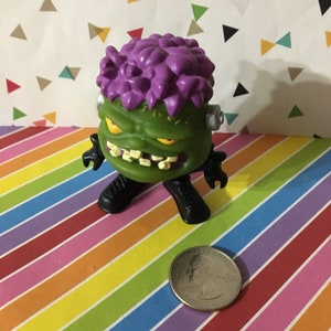May include: Green and purple monster toy with a toothy grin and black boots. The toy is about the size of a quarter.
