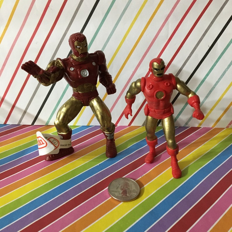 May include: Two red and gold Iron Man action figures. The figures are posed in a standing position. The figures are on a rainbow striped background.