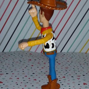 May include: A plastic toy figure of Woody from the Toy Story franchise. Woody is wearing his signature brown cowboy hat, yellow shirt, and blue jeans. He is holding his right arm up in the air, as if he is waving.