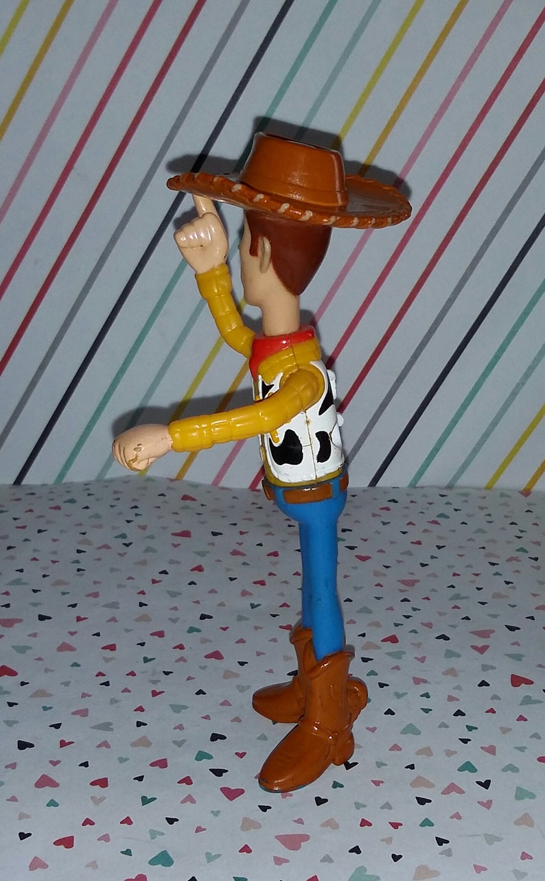 May include: A plastic toy figure of Woody from the Toy Story franchise. Woody is wearing his signature brown cowboy hat, yellow shirt, and blue jeans. He is holding his right arm up in the air, as if he is waving.