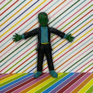 May include: A green Frankenstein's monster figurine with arms outstretched. The figure wears a black suit, a blue sweater, and blue shoes. The background features a colorful striped pattern.