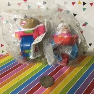 May include: Two plastic character toys packaged in clear plastic bags. The toys are blue, red, orange, pink, and green. The toys are shaped like a dog and a person. The dog is wearing a pink and white spacesuit. The person is wearing a pink and white dress. The toys are part of a Happy Meal promotion.