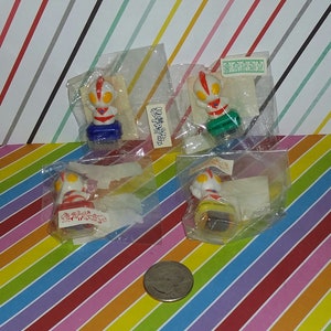 May include: Four small plastic figures of a superhero character in individual plastic bags. The figures are in different colors, including red, blue, green, and yellow. The bags have Japanese text on them.