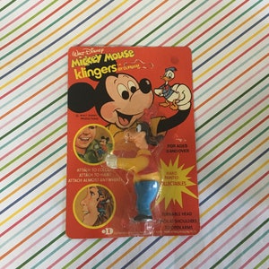May include: A vintage package for a Mickey Mouse Klinger toy. The package features a red background with a cartoon image of Mickey Mouse and Goofy. The text on the package reads "WALT DISNEY Mickey Mouse KLINGERS BY DURHAM". The package also includes instructions for attaching the toy to clothing or hair.