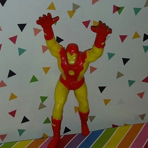 K&ouml;nnte beinhalten: Eine rote und gelbe Plastik-Actionfigur von Iron Man mit erhobenen Armen. Die Figur steht auf einer bunten gestreiften Oberfl&auml;che.