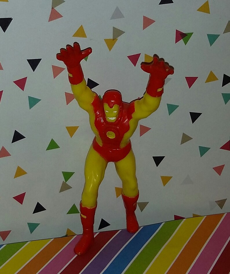 K&ouml;nnte beinhalten: Eine rote und gelbe Plastik-Actionfigur von Iron Man mit erhobenen Armen. Die Figur steht auf einer bunten gestreiften Oberfl&auml;che.