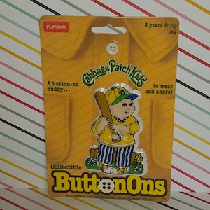May include: A yellow and green Cabbage Patch Kids button-on collectible. The collectible features a Cabbage Patch Kid wearing a yellow baseball uniform with a blue and white striped pants. The collectible includes a button for a "button-less" shirt. The packaging is yellow with the text "Cabbage Patch Kids" and "ButtonOns".