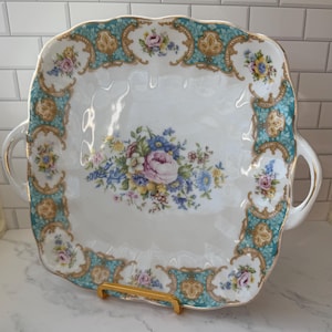 May include: Square white ceramic serving platter with a scalloped edge, adorned with a teal and gold border. The platter features a floral bouquet in the center and handles on each side. A gold-colored stand supports the platter.