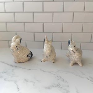 May include: Three ceramic rabbit figurines with a crackle glaze finish. The rabbits are off-white with black and blue accents. One rabbit is sitting, one is standing, and one is in a playful pose. The figurines are decorative and suitable for Easter.