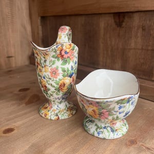 May include: A vintage floral creamer and sugar bowl set. The creamer has a handle and a flared spout. The sugar bowl is open with a gold rim. Both pieces are decorated with colorful flowers on a white background.