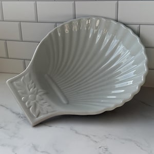 May include: A white, seashell-shaped ceramic dish with a ridged design. The dish has a decorative floral accent on the handle. The dish is on a white marble surface.