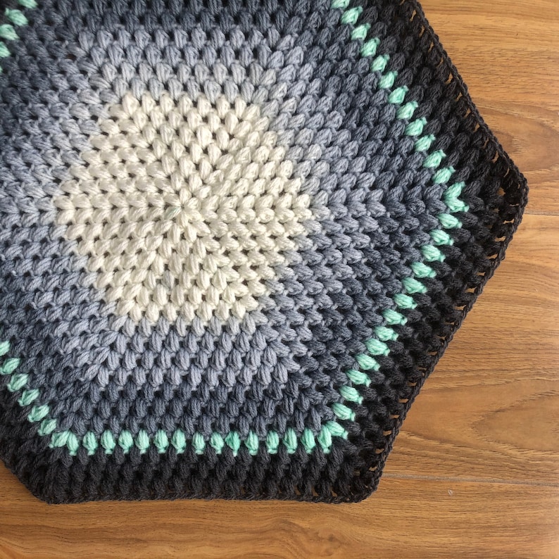 May include: A hexagonal crochet rug with a gradient of gray, white, and mint green colors. The rug has a black border and is made with a textured stitch.