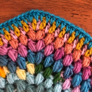 May include: A colorful crocheted blanket with a rainbow of colors, including blue, green, yellow, pink, and orange. The blanket is made with a textured stitch that creates a raised pattern.