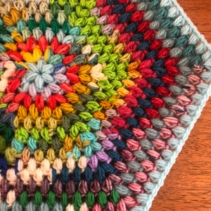 May include: A colorful crocheted square with a circular pattern in the center. The square is made with a variety of colors, including red, green, blue, yellow, and purple.