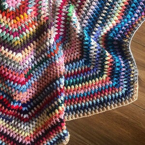 May include: A colorful crocheted blanket with a textured, bumpy surface. The blanket is made with a variety of different colored yarns, creating a patchwork effect. The blanket is folded in half and the edge is visible.