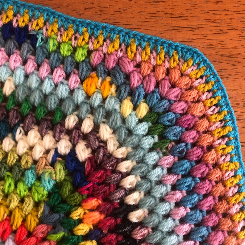 May include: A colorful crocheted blanket with a blue, yellow, and pink border. The blanket is made with a variety of different colored yarns.