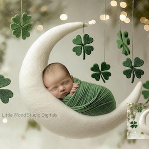 Könnte beinhalten: Ein Neugeborenes schläft auf einer weißen Halbmond-Requisite, eingewickelt in ein grünes Tuch. Der Mond hängt an einem Mobile mit grünen Kleeblatt-Dekorationen.
