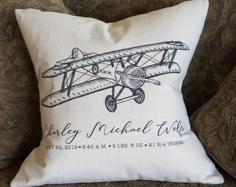 May include: A white pillow with a black and white illustration of a vintage airplane. The pillow has the text "Charley Michael Wolfe" and "May 20, 2019 - 9:46 A.M. - 9 LBS 8 OZ - 21 3/4 INCHES" printed on it.