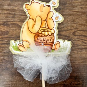 May include: A Winnie the Pooh cake topper with a yellow and brown bear holding a honey pot that says "Hunny". The topper is on a stick with a white tulle bow.